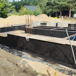 MTM-BUDOWA SPÓŁKA Z OGRANICZONĄ ODPOWIEDZIALNOŚCIĄ - Fundamenty domu dwulokalowego w trakcie izolacji przeciwwilgociowej, widoczne ściany fundamentowe zabezpieczone czarną powłoką, zbrojenie i częściowo ułożone bloczki.