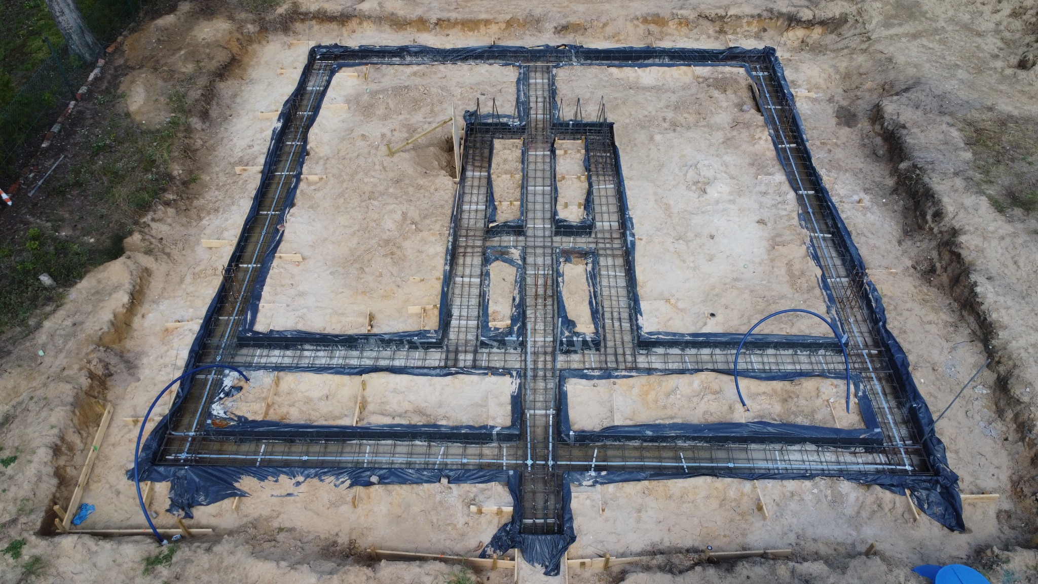 Zbrojenie ław fundamentowych pod budynek dwulokalowy, widok z góry. Zbrojenie w czarnej folii, częściowo zalane wodą. Wykopane fundamenty z piaskiem.