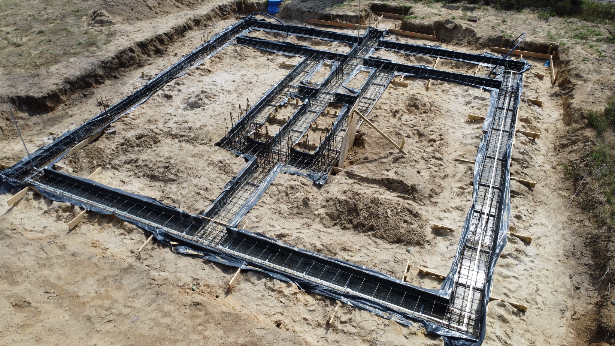 Zbrojenie ław fundamentowych domu dwulokalowego, widok z góry. Czarne folie pod zbrojeniem. Drewniane elementy szalunku. Piasek i ziemia wokół wykopu.