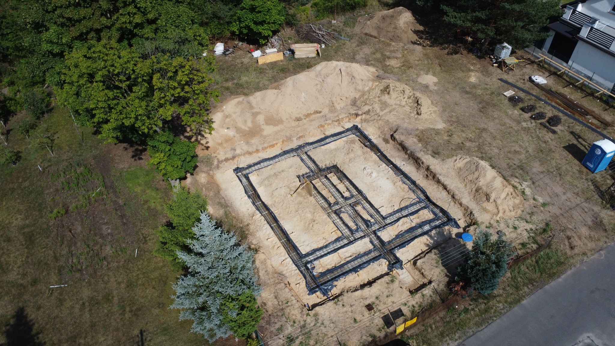 Zbrojenie ław fundamentowych pod budynek dwulokalowy, widok z drona. Czarna folia izolacyjna, wystające pręty zbrojeniowe. W tle drzewa i fragment budynku.
