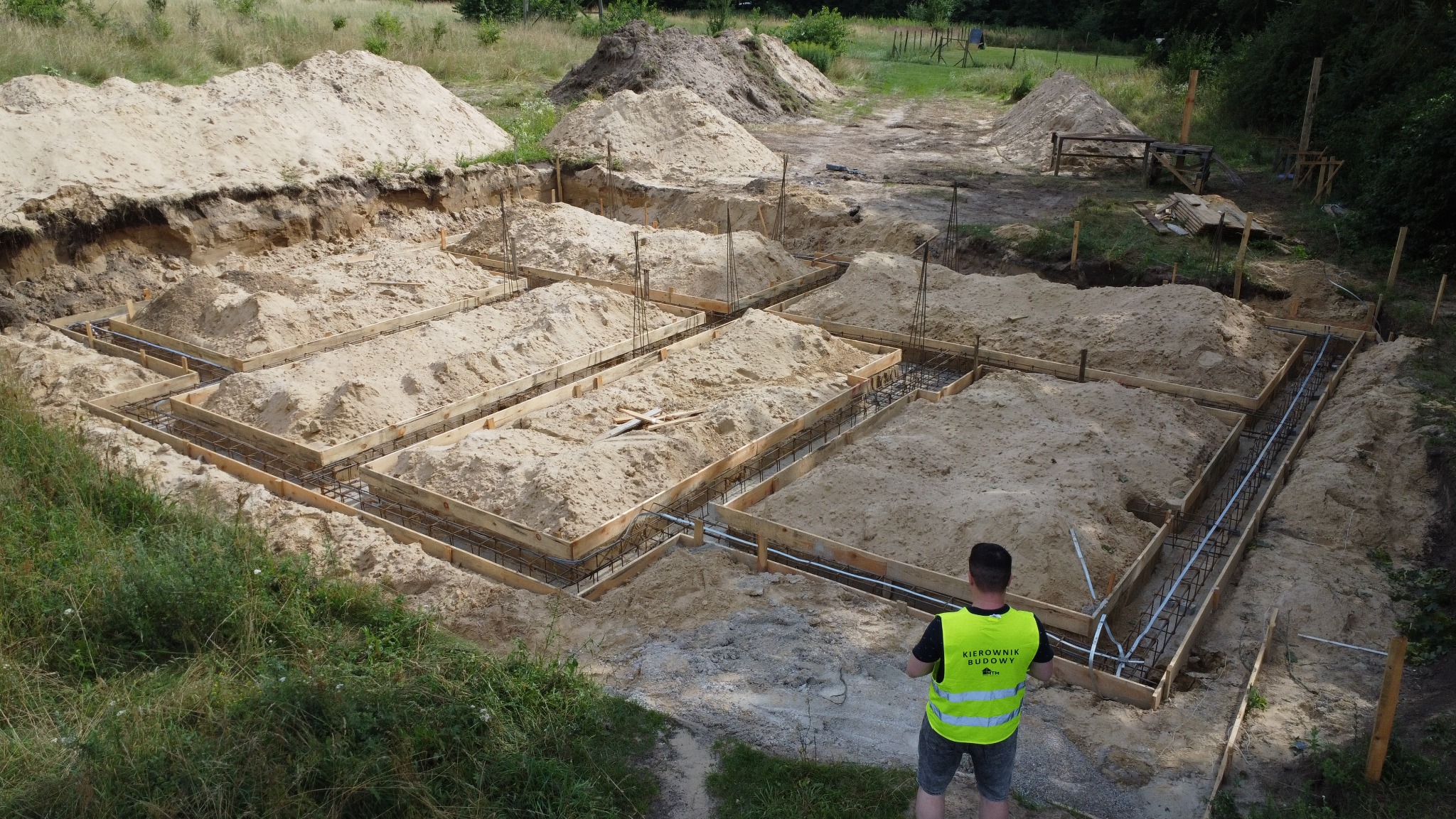 Ławy fundamentowe domu dwulokalowego w Pilchowie. Widoczne drewniane szalunki, zbrojenia i piasek. Kierownik budowy w kamizelce odblaskowej nadzoruje prace.