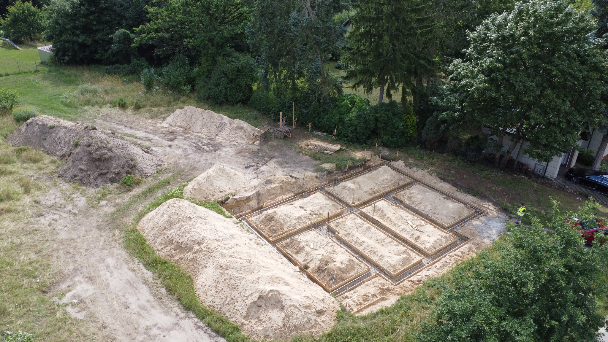 Widok z góry na plac budowy z wykopanymi i przygotowanymi ławami fundamentowymi pod budynek dwulokalowy, hałdy piachu, w tle zieleń i budynek.