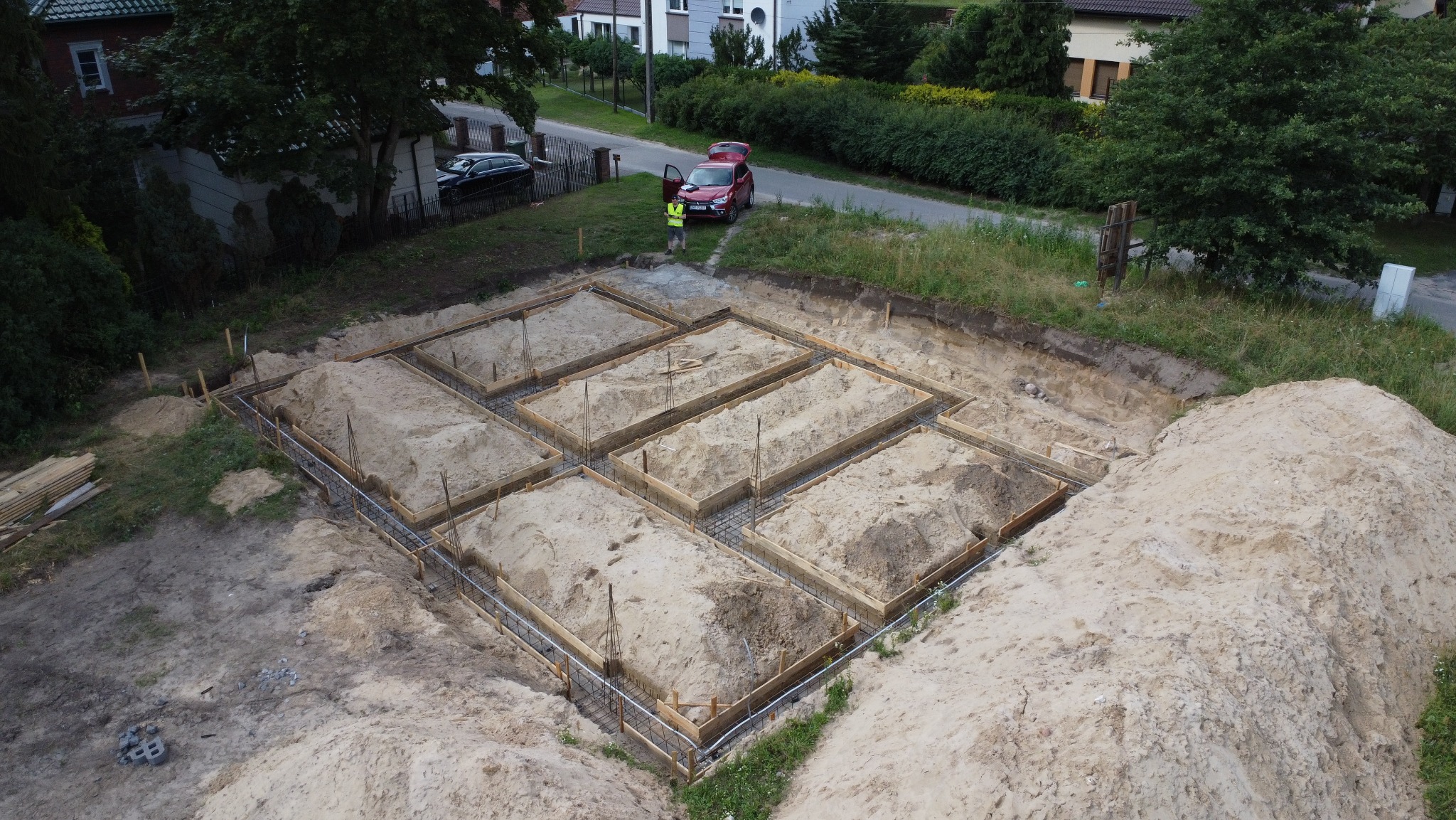 Budowa domu jednorodzinnego: widok z góry na wykopane i przygotowane ławy fundamentowe z drewnianym szalunkiem i zbrojeniem, obok stoi osoba w kamizelce.