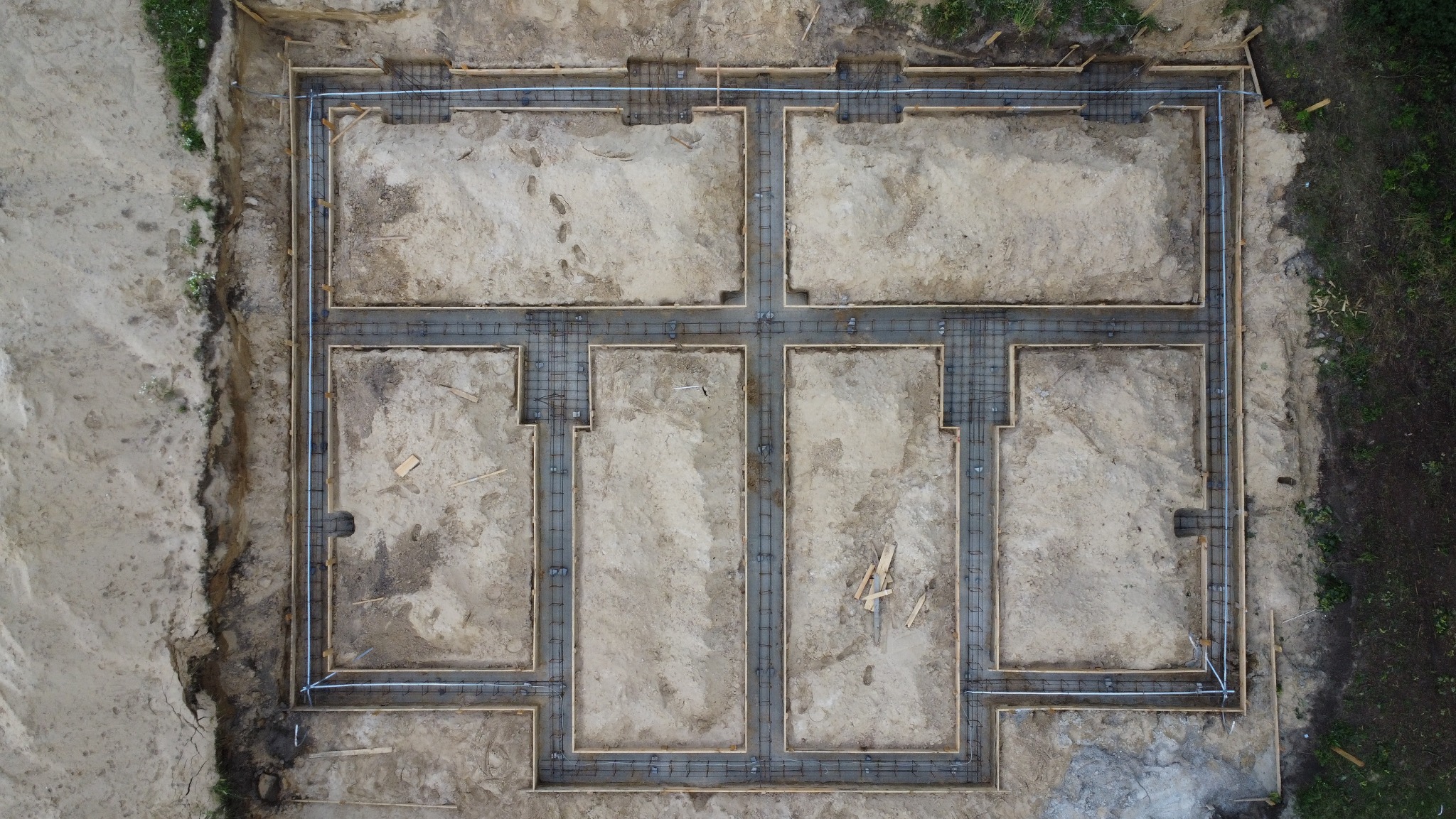 Widok z góry na wykopane i przygotowane ławy fundamentowe z zamontowanym zbrojeniem pod budowę domu dwulokalowego na piaszczystym terenie.