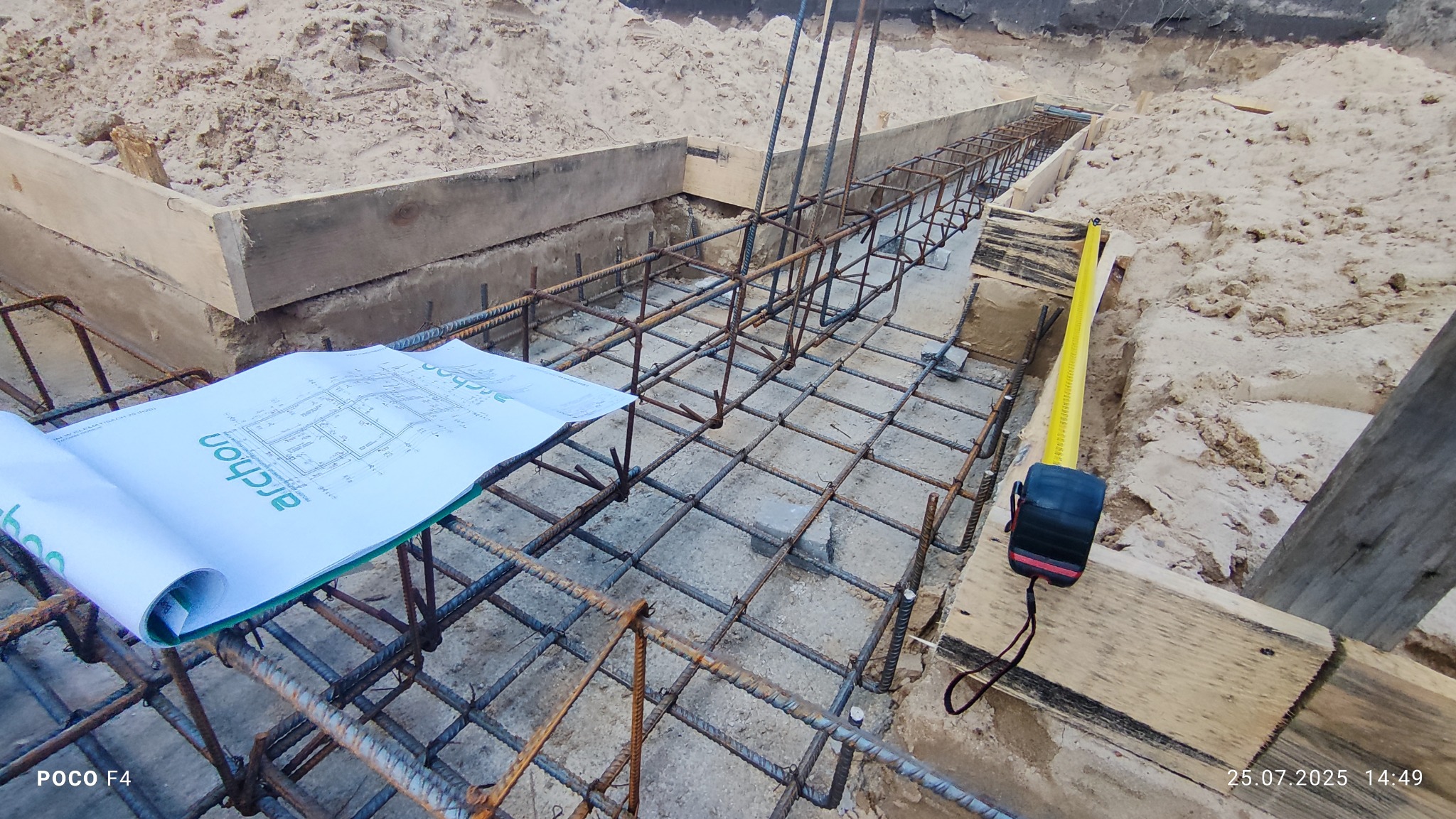 Zbrojenie ław fundamentowych na budowie domu jednorodzinnego, widoczne szalunki, plan architektoniczny i miara zwijana. Ujęcie z góry.