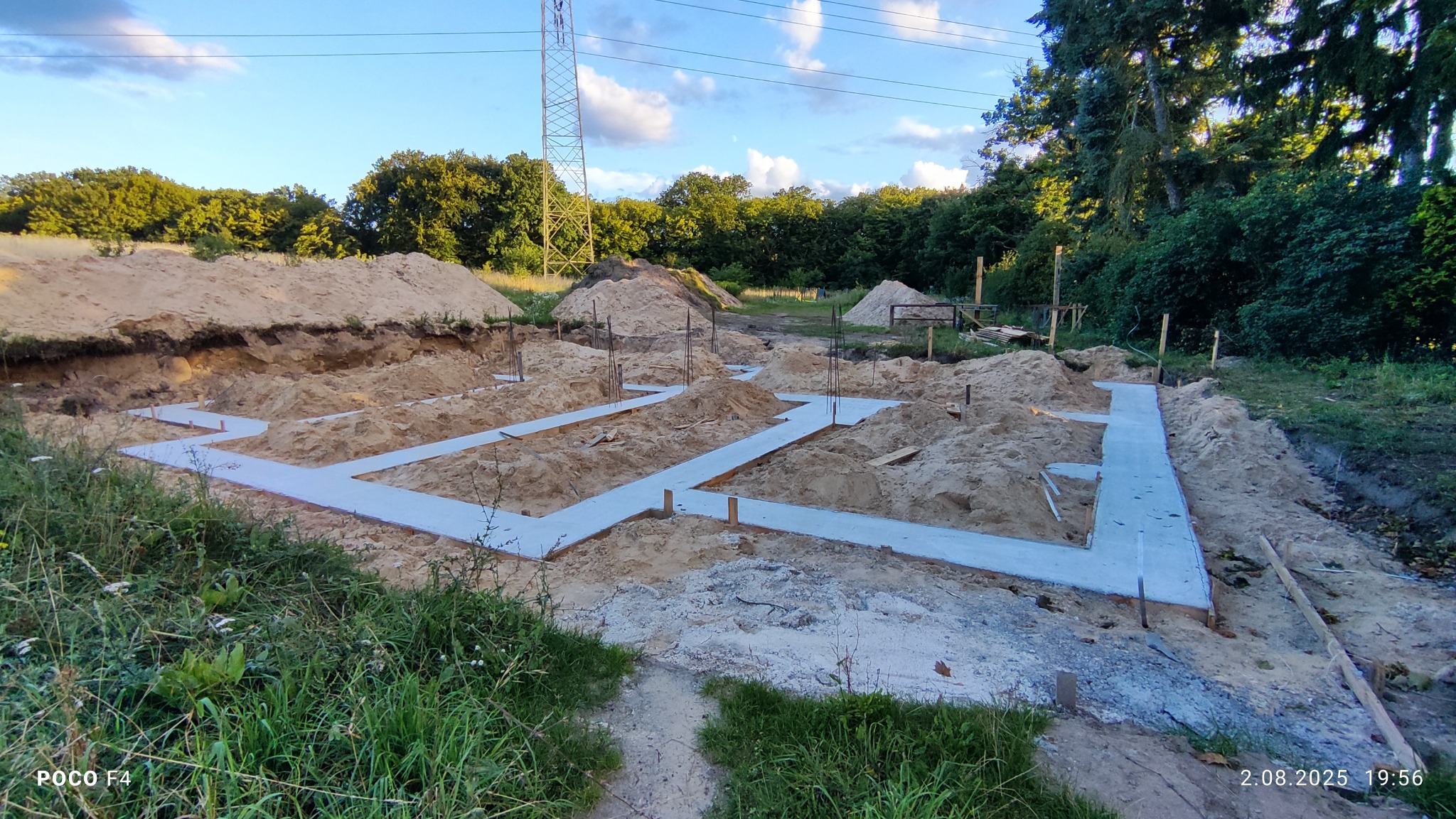 Budowa domu jednorodzinnego: wylane ławy fundamentowe w wykopie, widoczne wystające pręty zbrojeniowe. W tle hałda piasku i słup wysokiego napięcia.