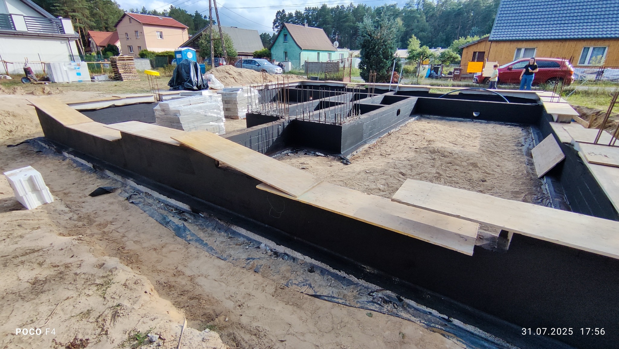 Fundamenty domu jednorodzinnego po izolacji przeciwwilgociowej, zbrojenie wewnątrz, widoczne deski szalunkowe i materiały budowlane w tle.