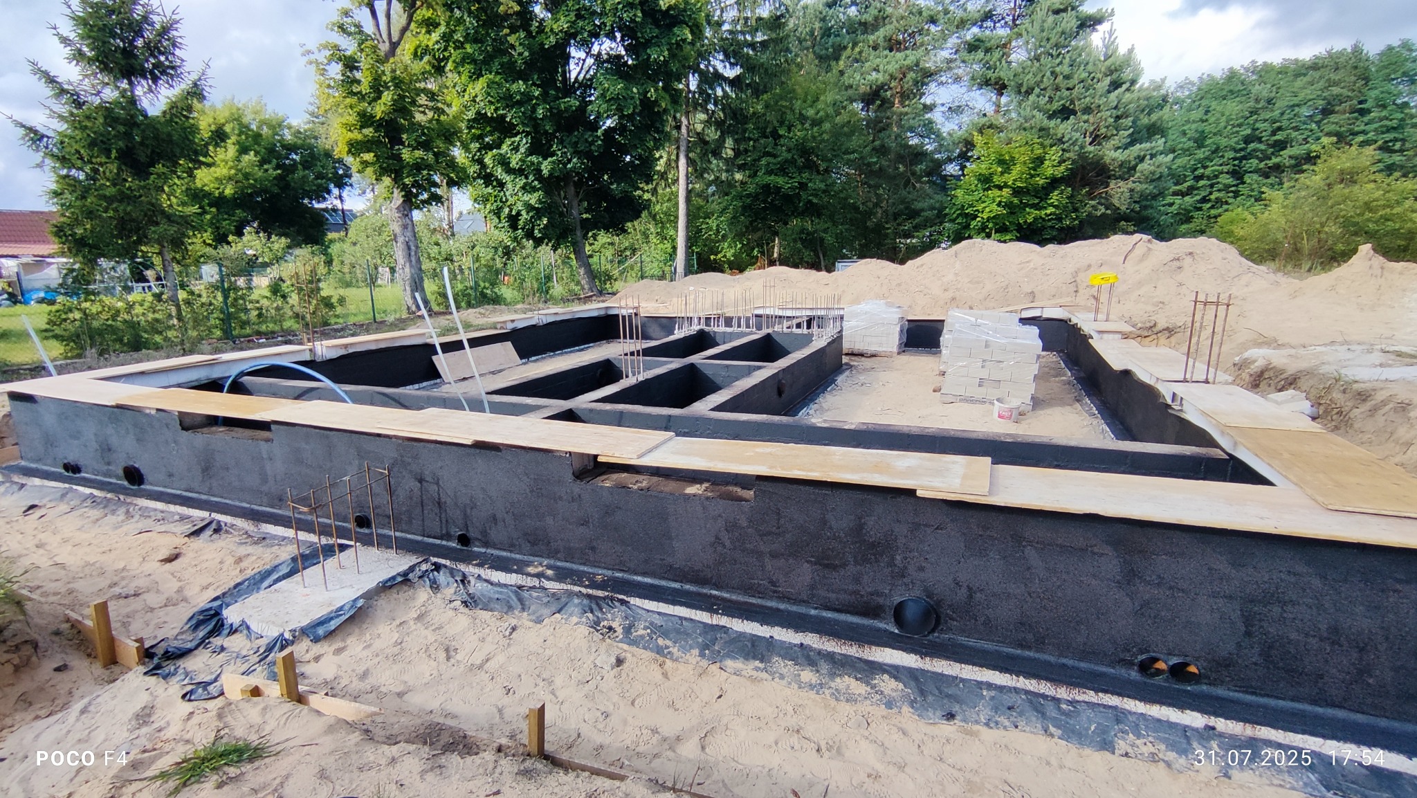 Fundamenty domu jednorodzinnego po izolacji przeciwwilgociowej, widoczne zbrojenia i przygotowane otwory wentylacyjne. Prace ziemne w toku.