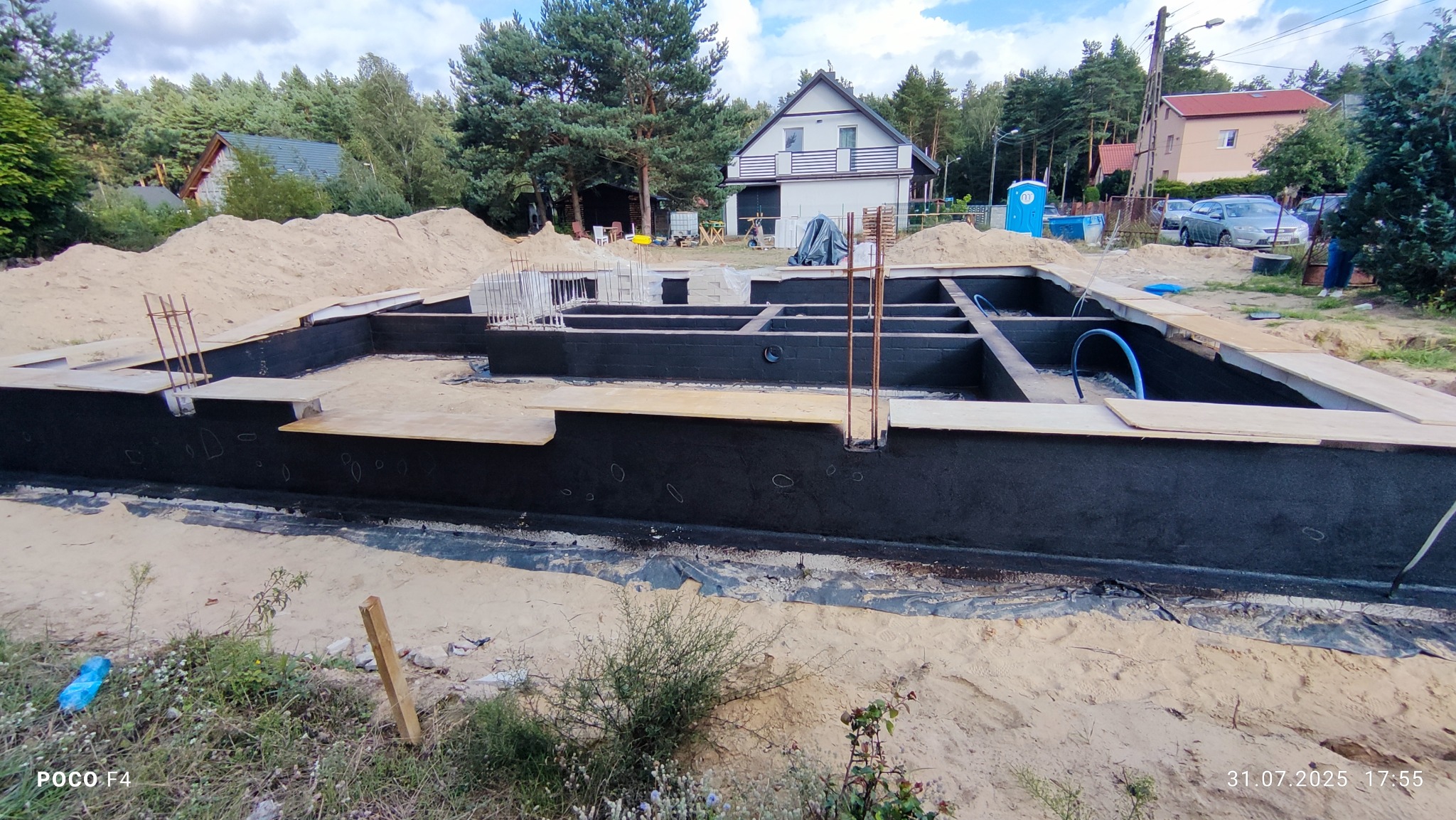 Izolacja przeciwwilgociowa fundamentów domu jednorodzinnego, widoczne czarne ściany fundamentowe, zbrojenie i przygotowany teren pod budowę.