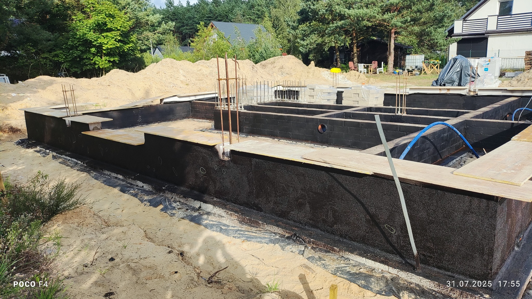 Fundamenty domu dwulokalowego w trakcie izolacji przeciwwilgociowej, widoczne ściany fundamentowe zabezpieczone czarną powłoką, zbrojenie i częściowo ułożone bloczki.