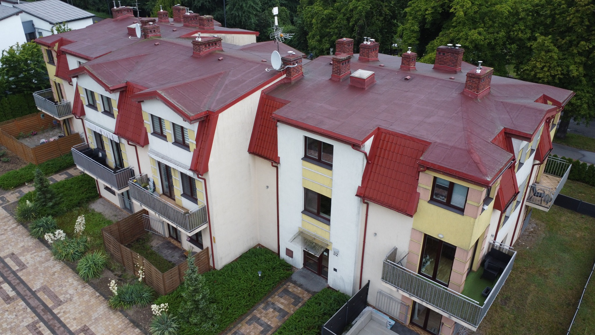 Wielorodzinny budynek z czerwonym dachem, widok z góry. Antena satelitarna na dachu. Balkony z szarymi barierkami. Uwagę zwraca zadbana zieleń wokół budynku.