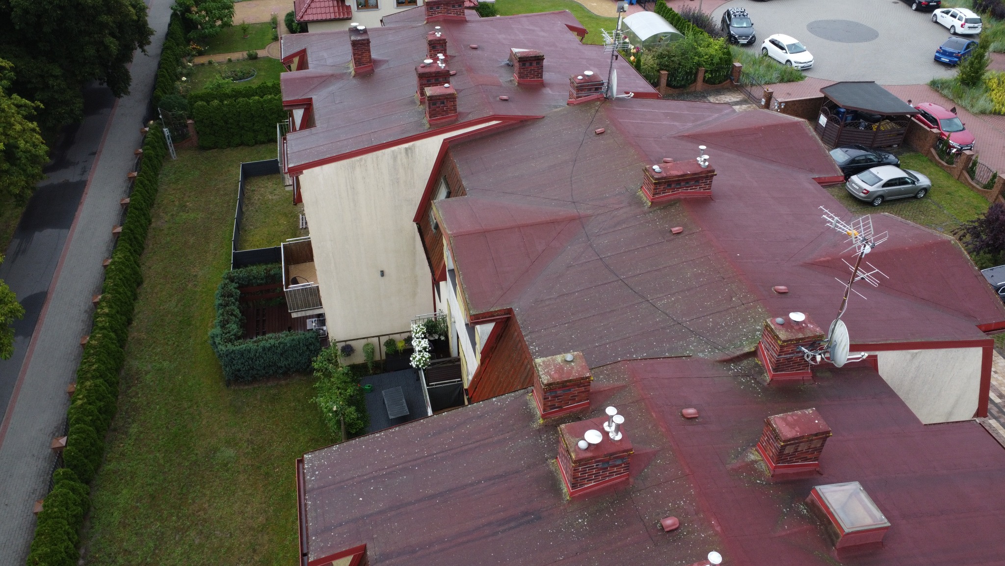 Widok z góry na dach budynku wielorodzinnego z kominami, anteną satelitarną i oknami dachowymi. Widoczny fragment osiedla z zielenią i zaparkowanymi autami.