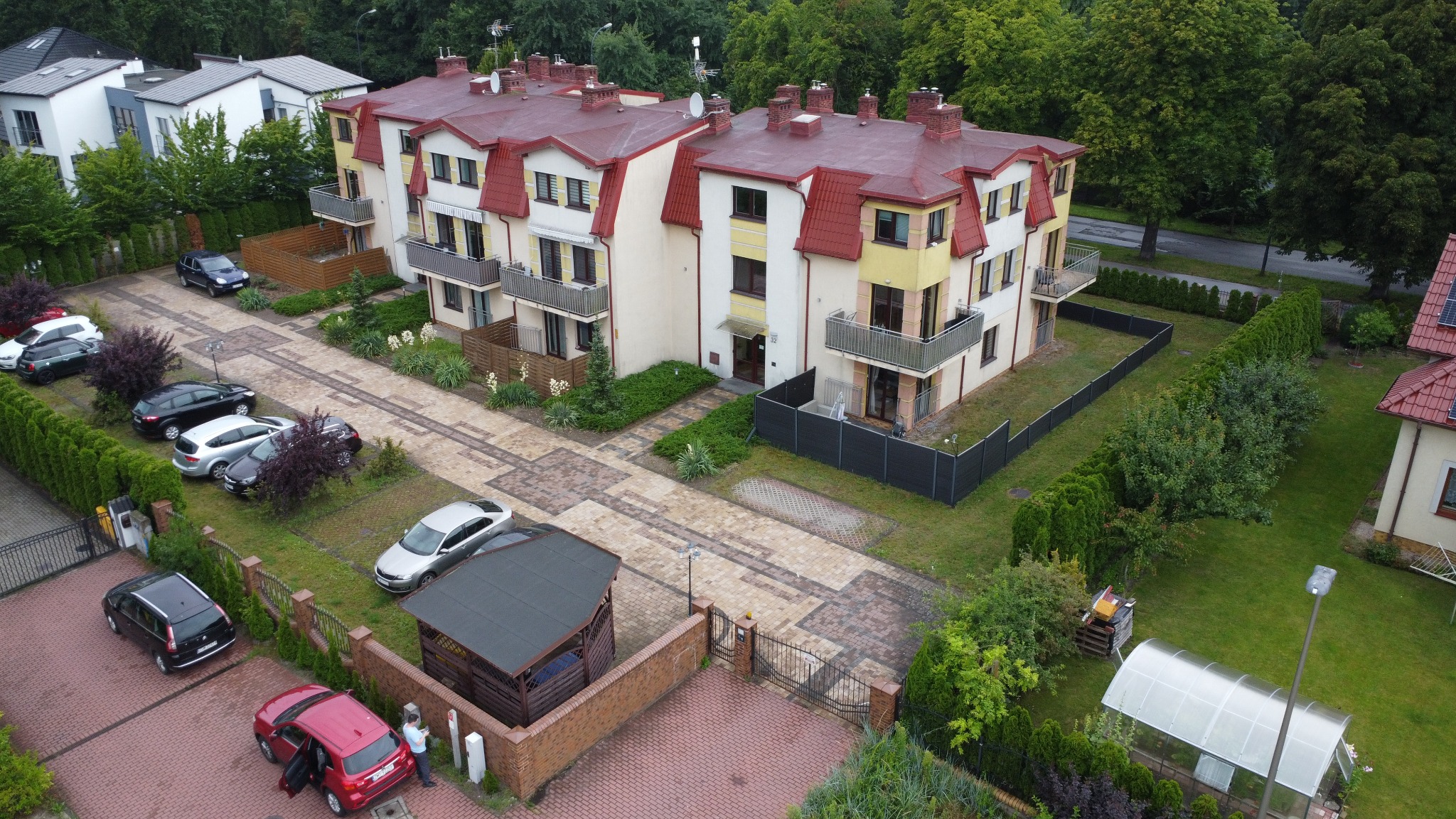 Wielorodzinny budynek z czerwonym dachem, widok z góry. Uwagę zwraca starannie zagospodarowany teren z parkingiem i zielenią. Widoczny fragment osiedla.