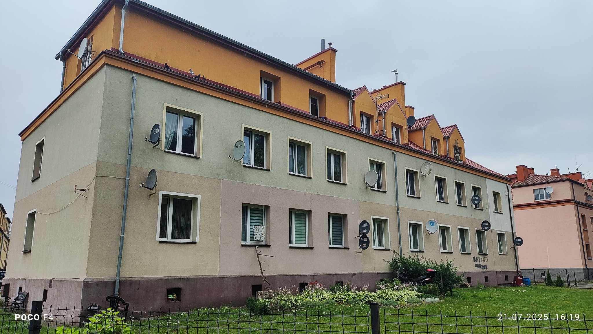 Wielorodzinny budynek z antenami satelitarnymi na elewacji, widoczny zza metalowego ogrodzenia. Budynek pomalowany w pasy beżu, szarości i żółci. Szary, pochmurny dzień.