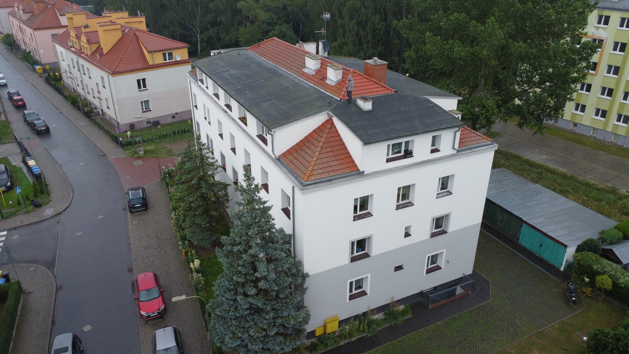 Wielorodzinny budynek z dachem częściowo pokrytym papą, częściowo czerwoną dachówką, widziany z góry. Na dachu kominy. Obok drzewa i ulica z samochodami.