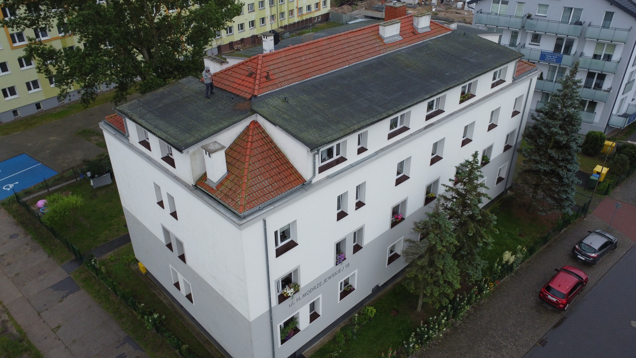Przegląd budynku wielorodzinnego z góry: inspektor na dachu ocenia stan techniczny. Widoczne okna z kwiatami, drzewa i zaparkowane auta.