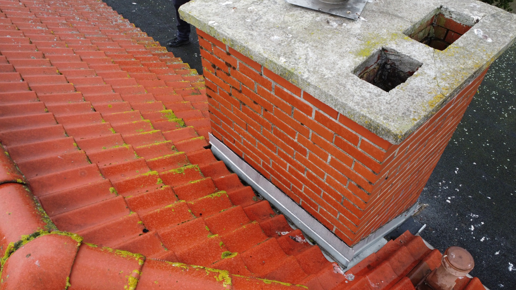 Ceglany komin z betonową koroną i dwoma otworami na dachu z czerwonej dachówki, częściowo pokrytej mchem. Widoczna metalowa obróbka komina.