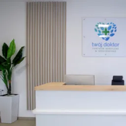 Recepcja w centrum medycznym z drewnianym blatem, białym frontem, monitorem komputera, czarnym krzesłem, logotypem 'Twój Doktor' na ścianie i rośliną doniczkową w białej donicy.