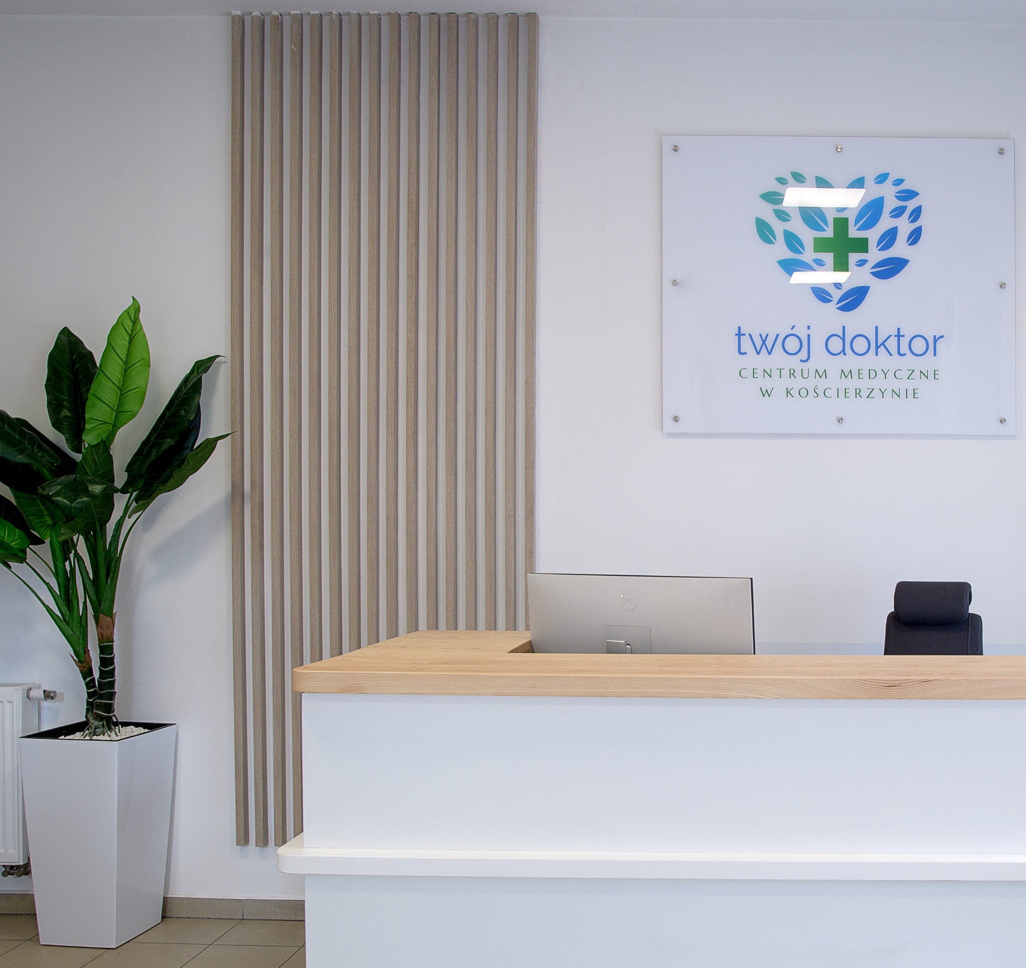 Recepcja w centrum medycznym z drewnianym blatem, białym frontem, monitorem komputera, czarnym krzesłem, logotypem 'Twój Doktor' na ścianie i rośliną doniczkową w białej donicy.