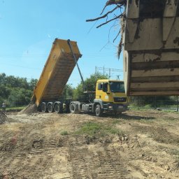 KRBM Mateusz Łabuziński - Żółta wywrotka MAN wysypuje ziemię na plac budowy, w tle zieleń i błękitne niebo. Łyżka koparki z ziemią w prawym górnym rogu kadru.
