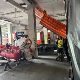KRBM Mateusz Łabuziński - Wnętrze remizy strażackiej: wywrotka z podniesioną skrzynią, pracownik w kamizelce, łódź ratunkowa na przyczepie. W tle wozy strażackie i instalacje pod sufitem.