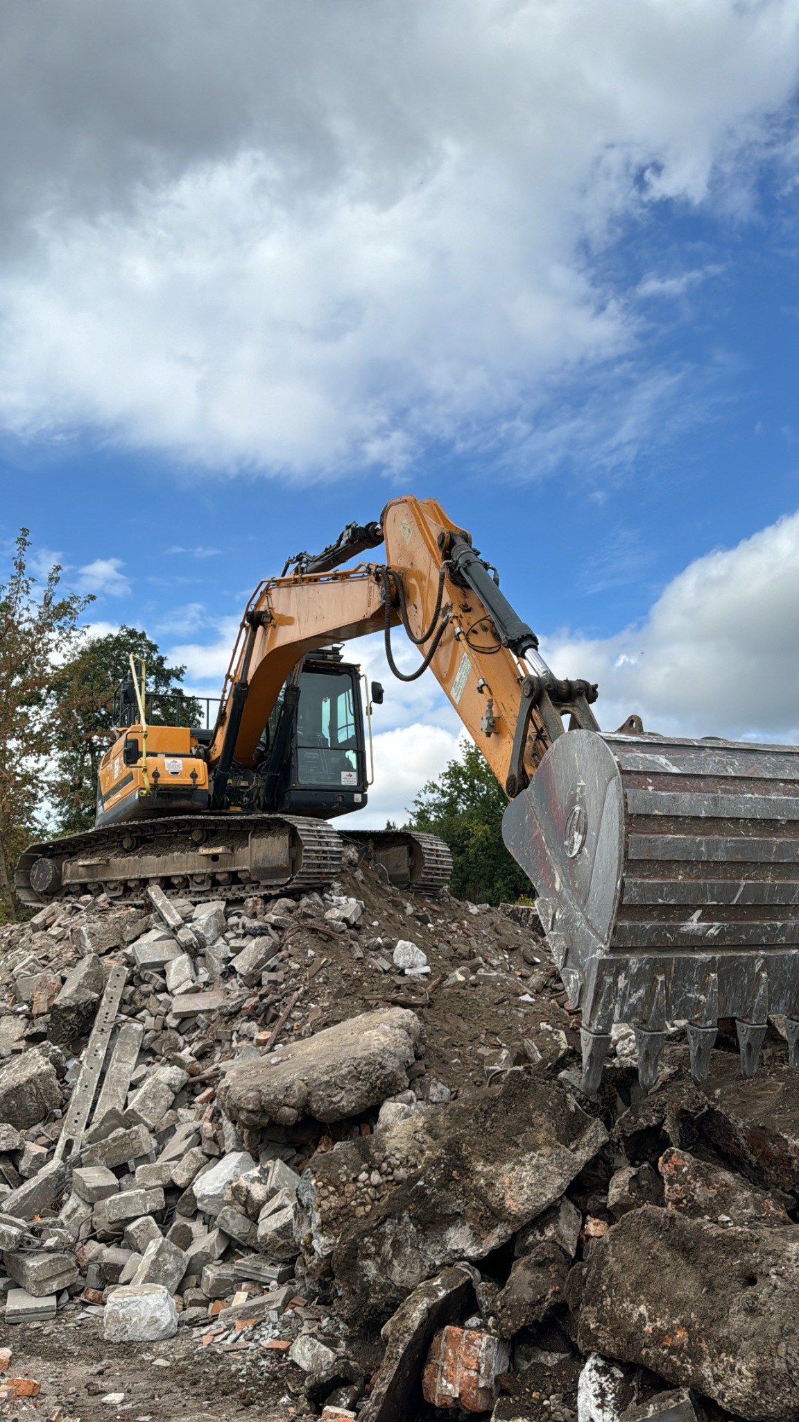 Żółta koparka gąsienicowa stoi na gruzowisku z betonu i cegieł na tle błękitnego nieba z chmurami, widoczne drzewa w tle.