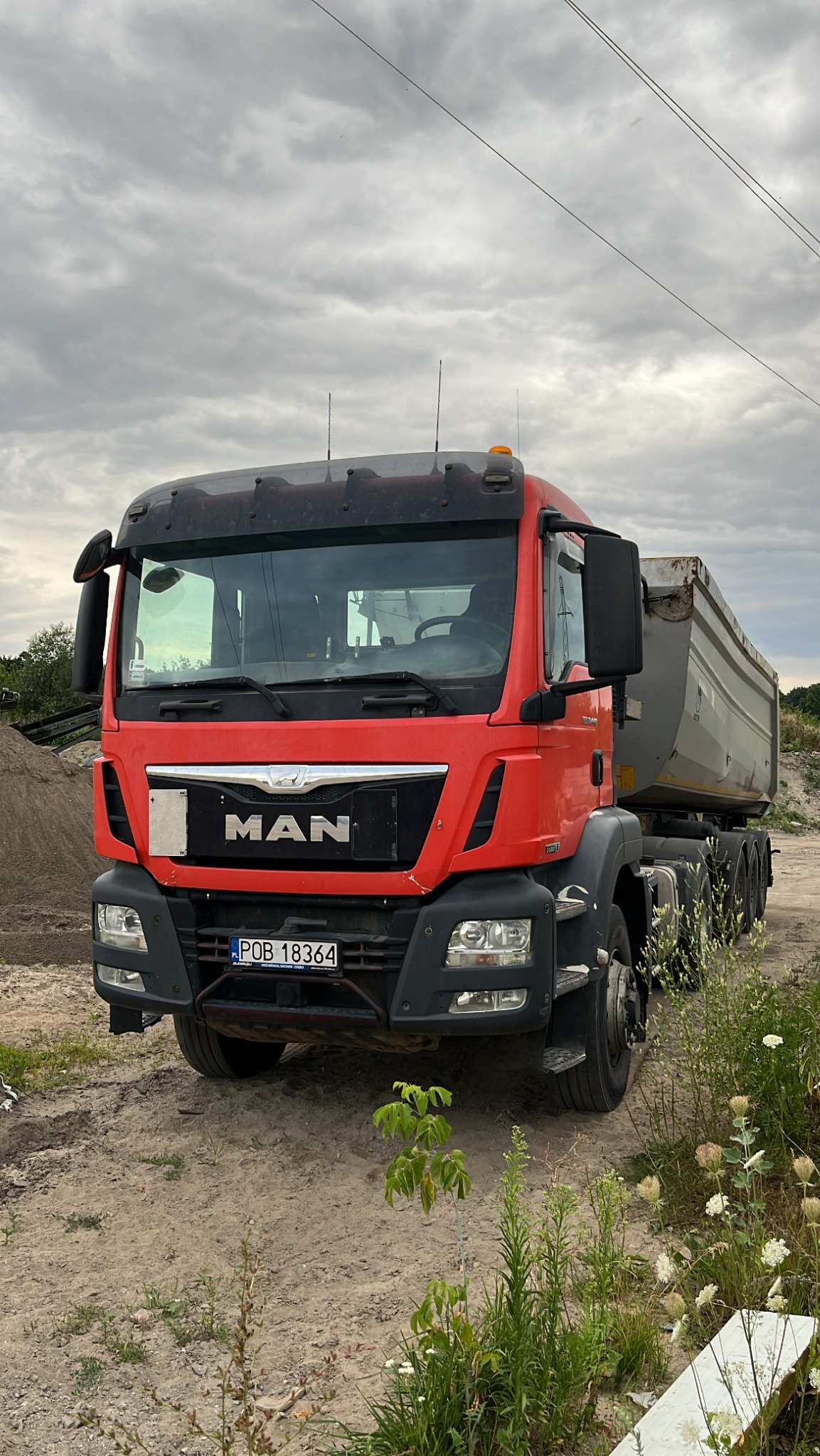 Czerwona wywrotka MAN zaparkowana na placu budowy, z widoczną stertą piasku w tle i młodymi drzewkami na pierwszym planie, pochmurne niebo.