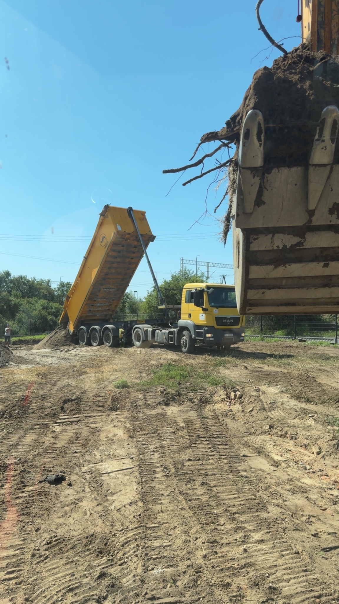 Żółta wywrotka MAN wysypuje ziemię na plac budowy, w tle zieleń i błękitne niebo. Łyżka koparki z ziemią w prawym górnym rogu kadru.