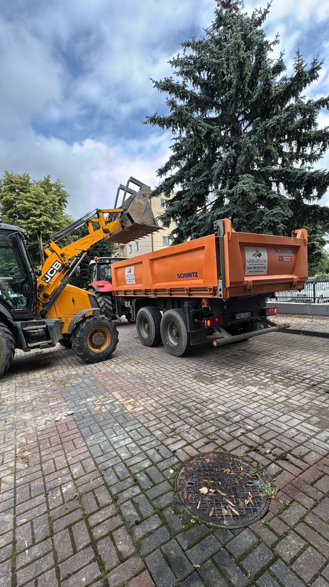 Żółta koparko-ładowarka JCB ładuje pomarańczową wywrotkę Schmitz na placu z kostki brukowej. W tle drzewa i budynek. Prace ziemne.