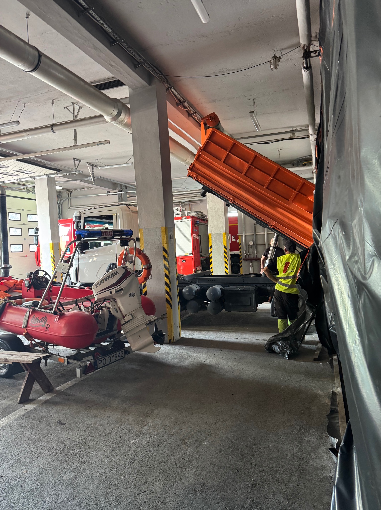 Wnętrze remizy strażackiej: wywrotka z podniesioną skrzynią, pracownik w kamizelce, łódź ratunkowa na przyczepie. W tle wozy strażackie i instalacje pod sufitem.