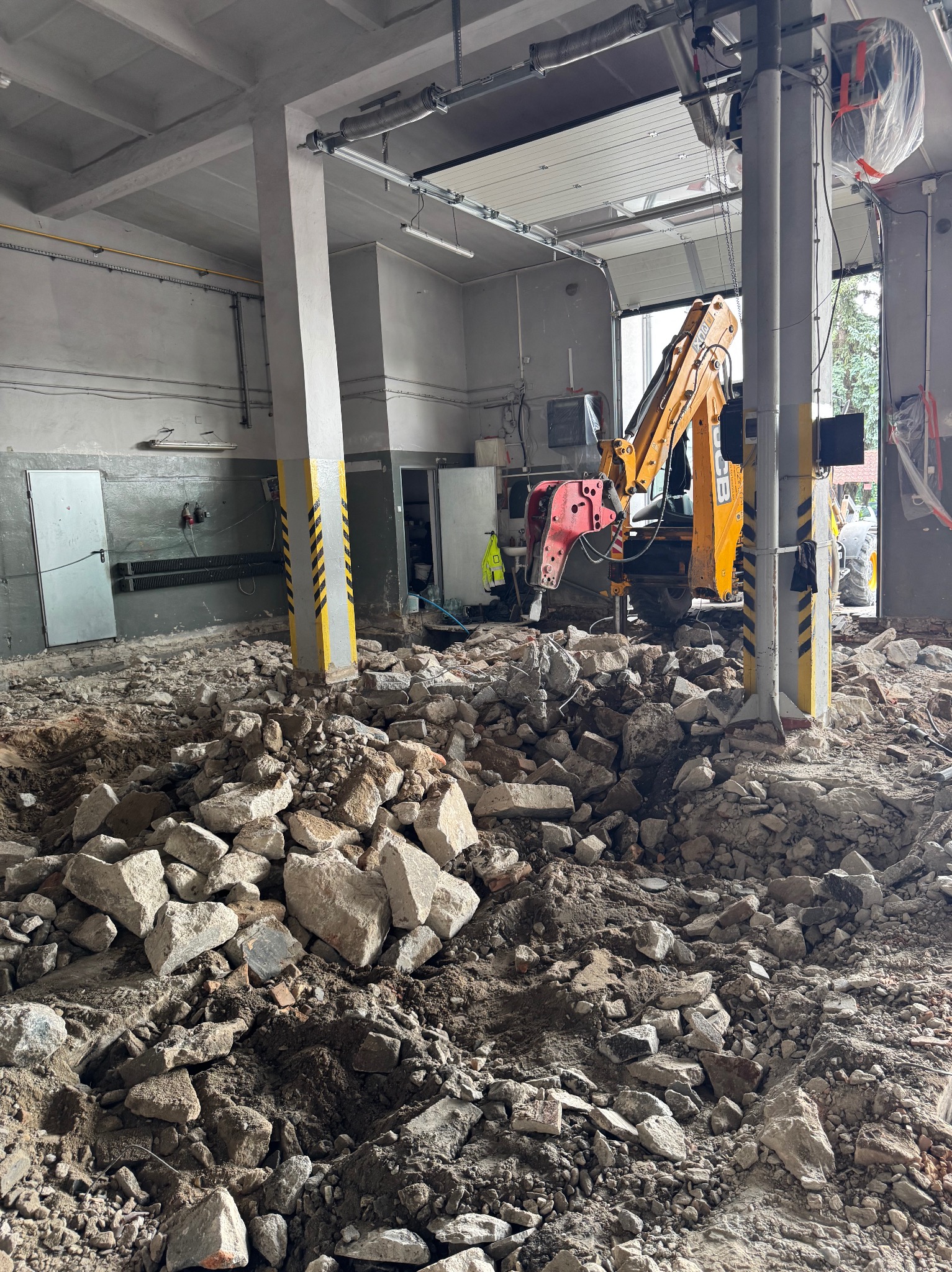 Wnętrze budynku straży pożarnej w trakcie prac rozbiórkowych: gruz, wykopany grunt i koparko-ładowarka JCB z zamontowanym młotem pneumatycznym.