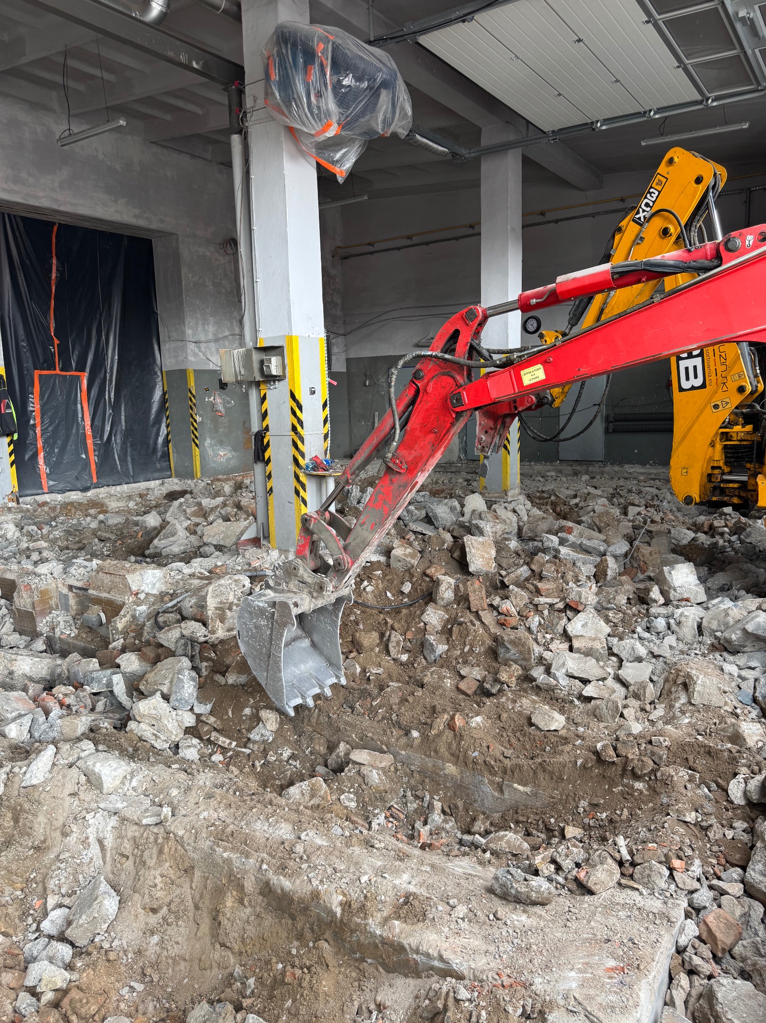 Czerwono-żółta koparka w trakcie prac rozbiórkowych wewnątrz budynku. Gruz i wykopana ziemia na podłożu, widoczne filary i elementy konstrukcyjne.