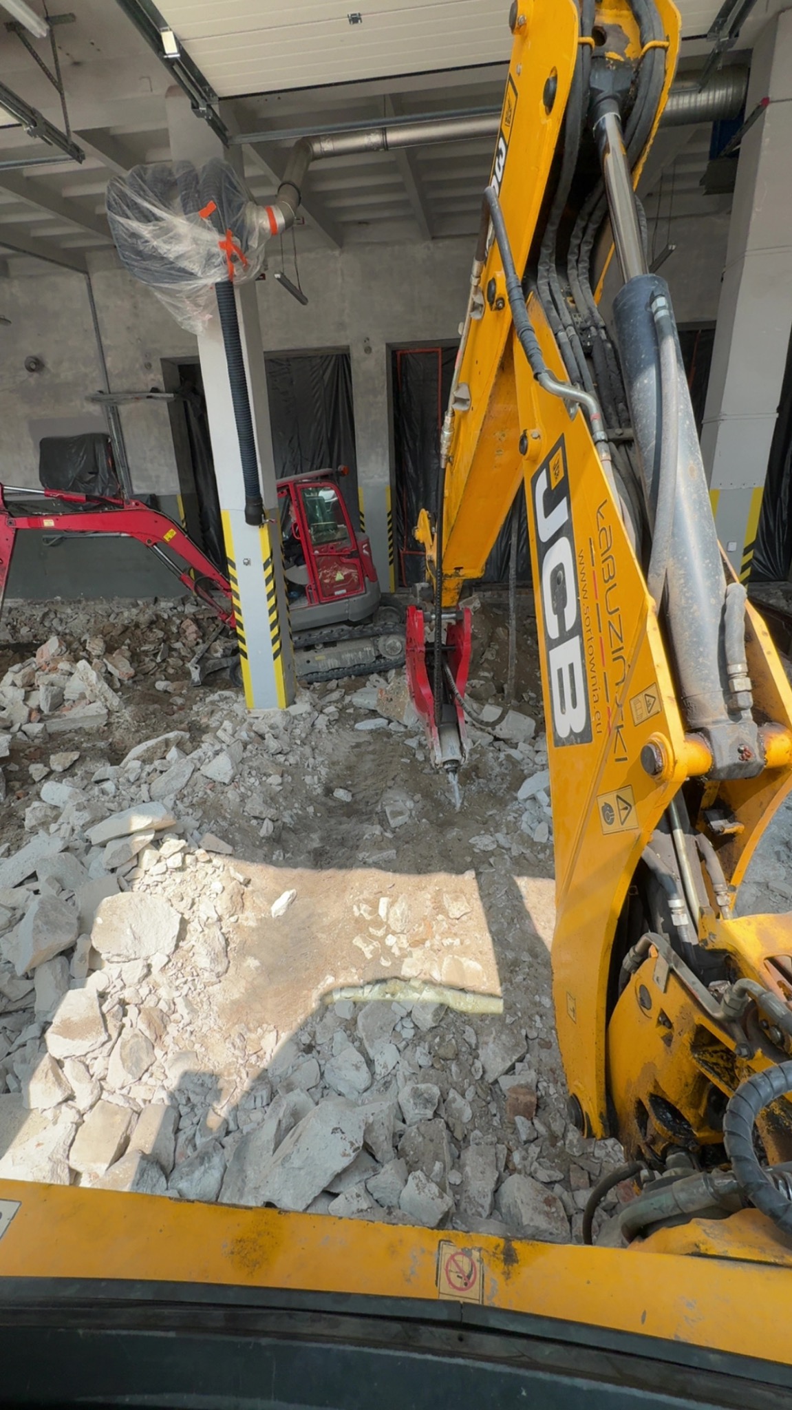 Żółty młot hydrauliczny JCB kruszy gruz wewnątrz budynku, w tle widoczny czerwony minikoparka. Prace rozbiórkowe w hali.