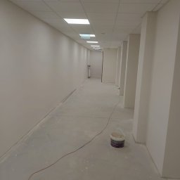 BUD-MASTER YURII ZAKHARENKO - Długi korytarz biurowy po malowaniu na jasny kolor, z widocznym wiadrem farby przy filarze. Sufit podwieszany z oświetleniem. Świeżo pomalowane ściany i posadzka.