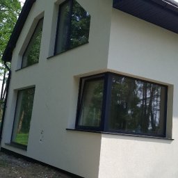 BUD-MASTER - Ukończona elewacja nowoczesnego domu w jasnym kolorze, z ciemnym dachem i ramami okiennymi. Charakterystyczne trójkątne okna szczytowe i duże okno narożne.