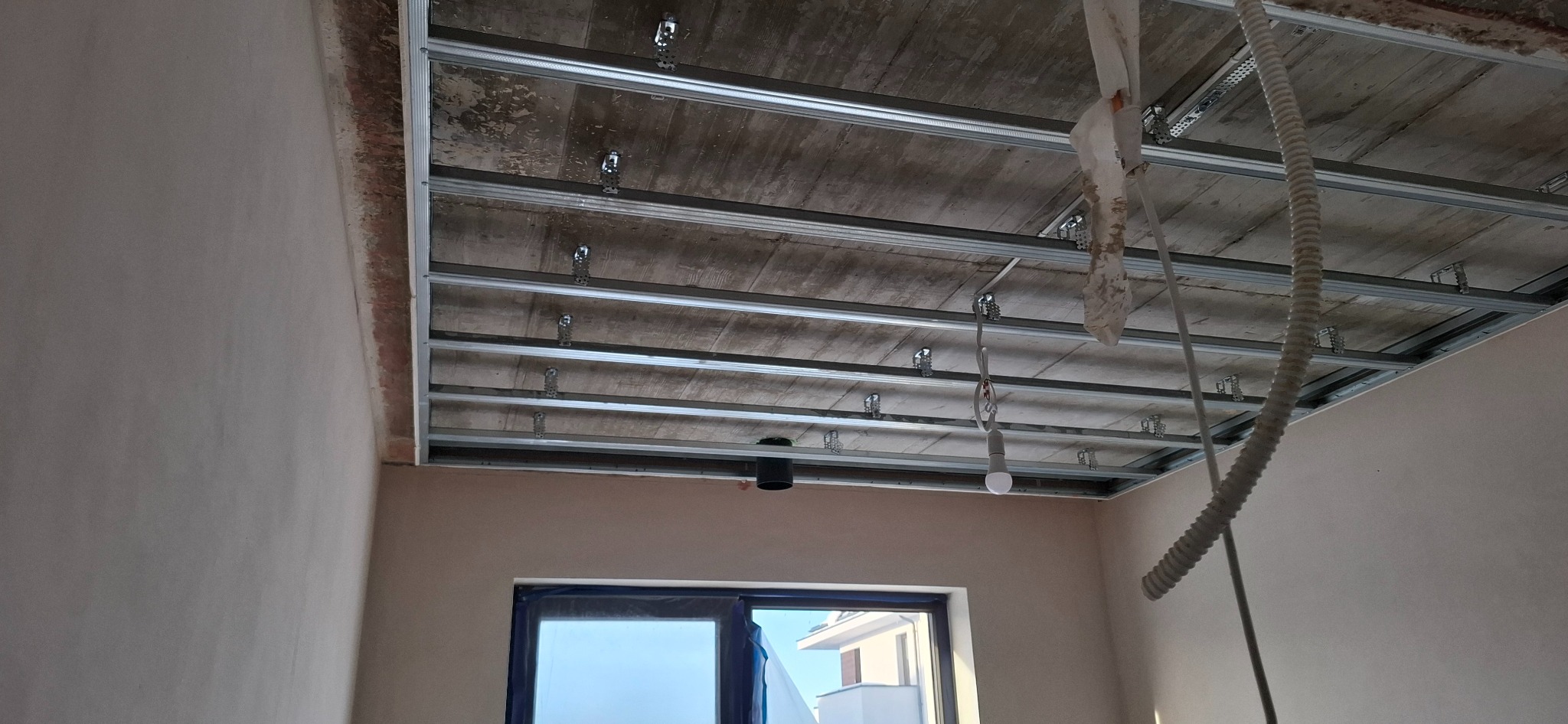 Sufit z metalową konstrukcją podwieszaną, widoczne profile i uchwyty montażowe. W tle ściany w jasnym odcieniu i okno zabezpieczone folią. Etap prac wykończeniowych.