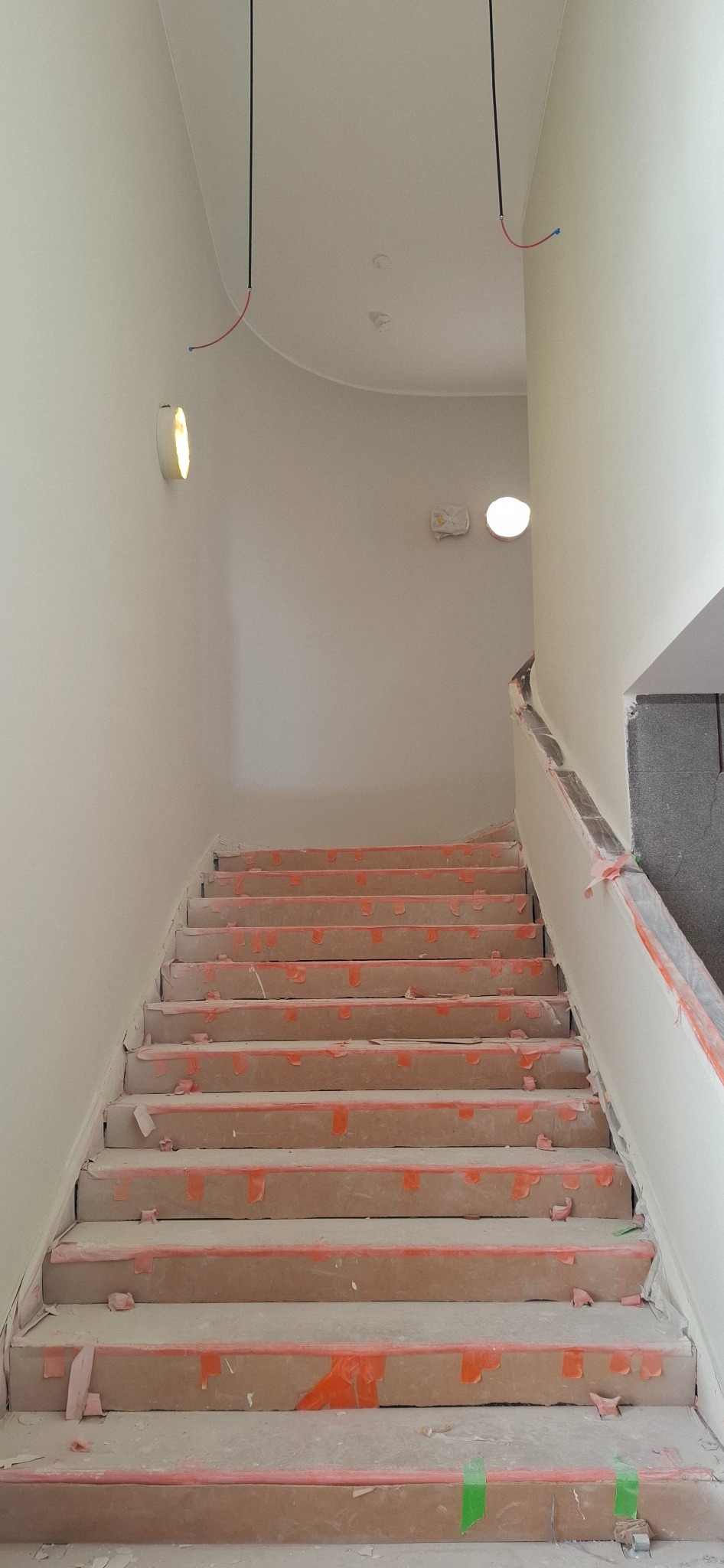 Klatka schodowa w trakcie prac wykończeniowych: zabezpieczone schody, świeżo pomalowane ściany, wystające przewody elektryczne. Widok z dołu na górę.
