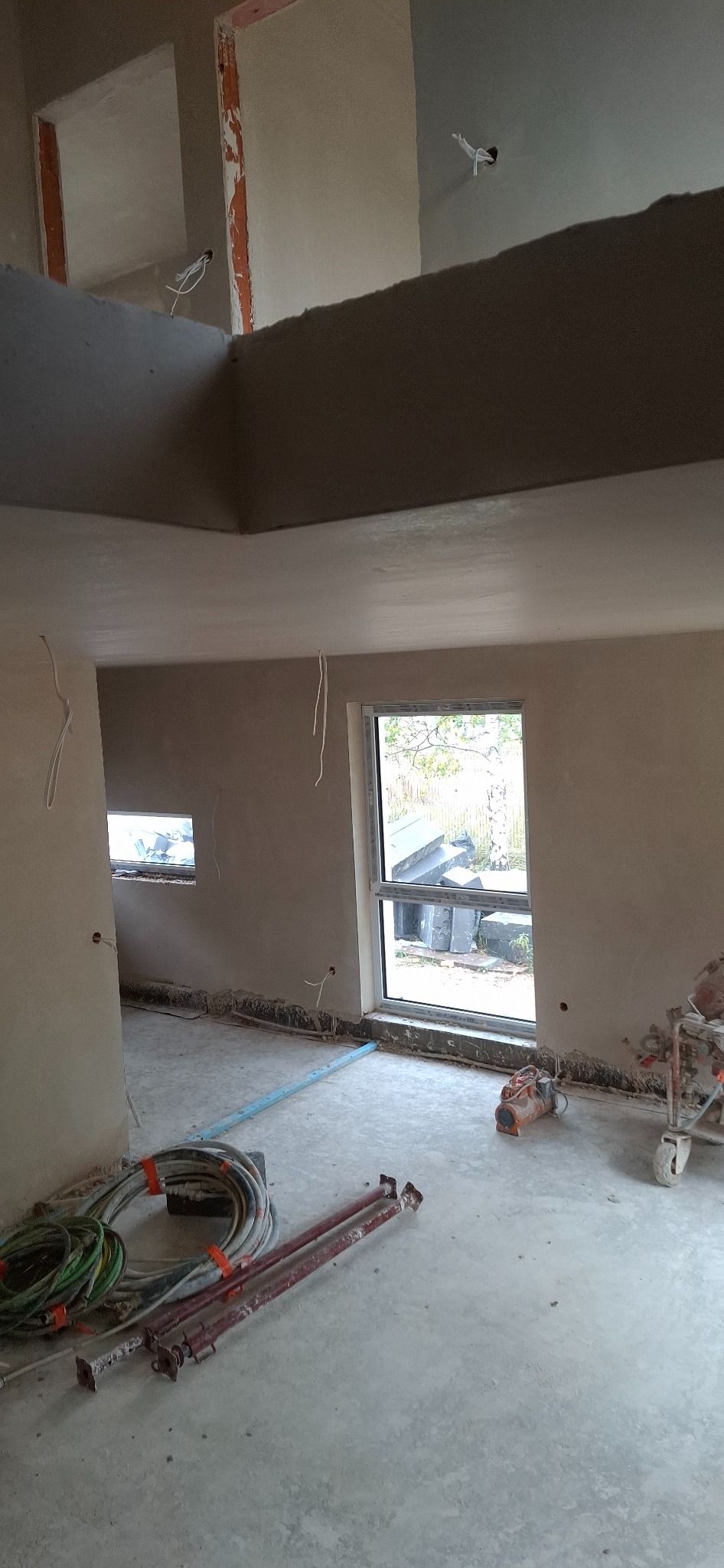 Wnętrze w trakcie prac wykończeniowych: świeżo położone tynki maszynowe na ścianach i suficie, widoczne przewody elektryczne, okno z widokiem na zewnątrz i narzędzia pracy.