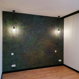 Kamil Grącki - Pokój po odświeżeniu: ściana z efektem patyny w odcieniach zieleni i brązu, dwie wiszące lampy w stylu industrialnym, parkiet w kolorze jasnego drewna.