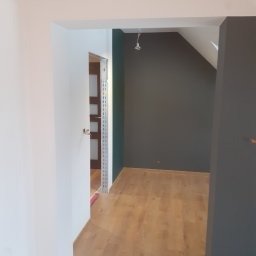 Kamil Grącki - Wnętrze w trakcie odświeżania: pomalowana na szaro ściana w pokoju na poddaszu, widok z sąsiedniego pomieszczenia z białymi ścianami i otwartymi drzwiami, na podłodze panele w odcieniach brązu.