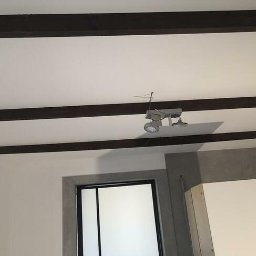 Kamil Grącki - Wnętrze pokoju z białymi ścianami, ciemnymi belkami na suficie, szarą wnęką i nowoczesnym oświetleniem punktowym