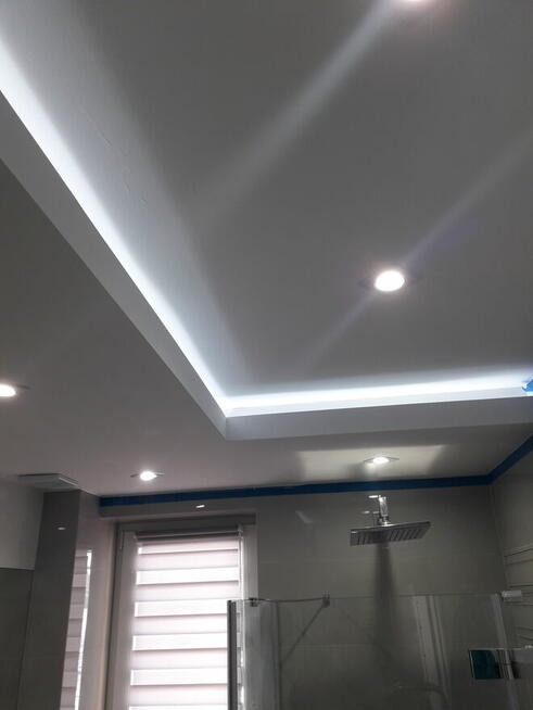 Nowoczesna łazienka z podwieszanym sufitem z oświetleniem LED, szklaną kabiną prysznicową i roletami w oknie.