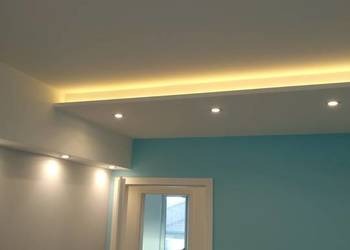 Turkusowa ściana z białym sufitem podwieszanym, oświetlonym listwą LED i trzema punktowymi lampami halogenowymi, widoczne białe drzwi.