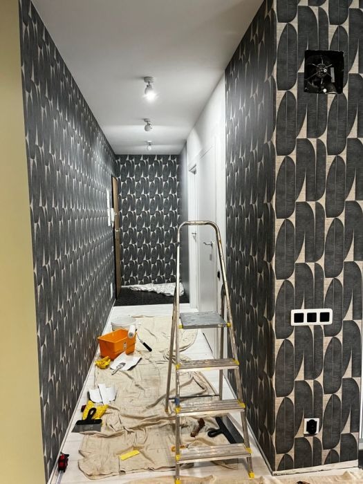 Korytarz w trakcie tapetowania z użyciem drabiny, narzędzi malarskich i wzorzystej tapety w odcieniach szarości i beżu.