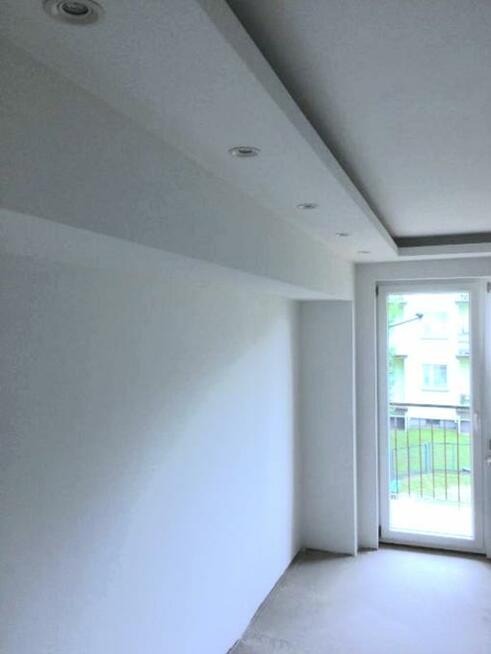 Pomalowane na biało wnętrze z sufitowym oświetleniem punktowym i oknem balkonowym, widok na zewnątrz.