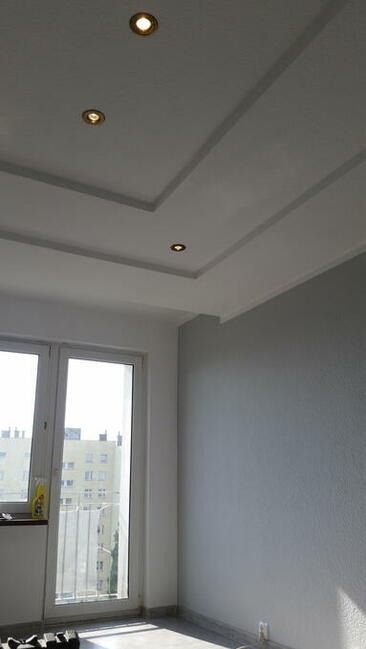 Pomieszczenie po remoncie: szara ściana, biały sufit z dekoracyjnymi listwami i trzema wbudowanymi lampami, okno balkonowe z widokiem na budynek mieszkalny.