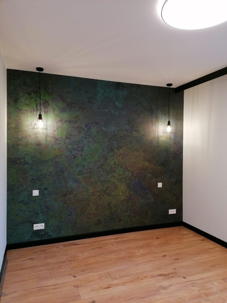Pokój po odświeżeniu: ściana z efektem patyny w odcieniach zieleni i brązu, dwie wiszące lampy w stylu industrialnym, parkiet w kolorze jasnego drewna.
