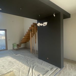 Wnętrze w trakcie remontu: pomalowany na ciemno sufit i fragment ściany, drewniane schody bez balustrady, zabezpieczona folią podłoga, widoczne ślady po szpachlowaniu na ścianie.