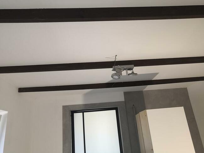 Wnętrze pokoju z białymi ścianami, ciemnymi belkami na suficie, szarą wnęką i nowoczesnym oświetleniem punktowym