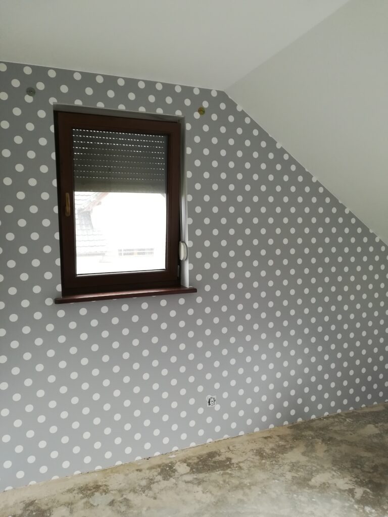 Pokój na poddaszu po malowaniu ścian na szaro w białe kropki, widoczne okno z roletą i surowa podłoga.
